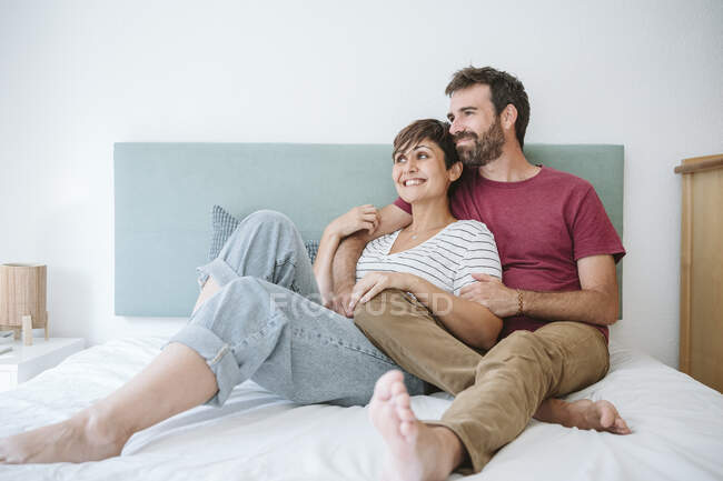 Glückliches erwachsenes Paar, das wegschaut, während es zu Hause zusammen auf dem Bett sitzt — Stockfoto