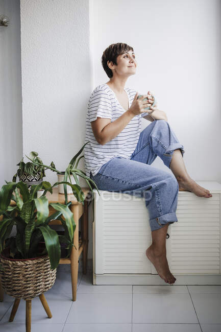 Mujer adulta con taza sentada cerca de las plantas en casa - foto de stock