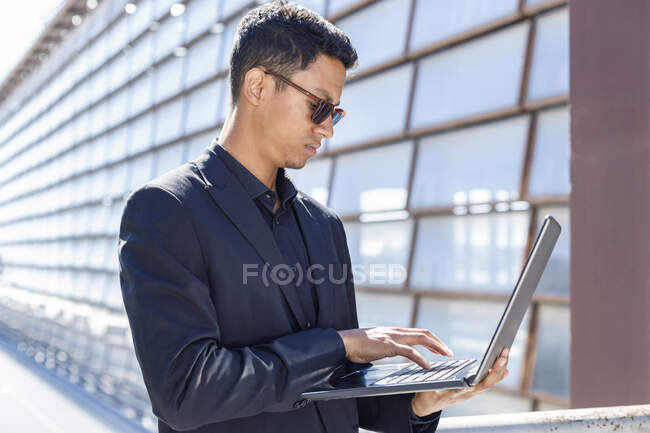 Профессиональная мужская печать на ноутбуке, стоя снаружи офисного здания — стоковое фото