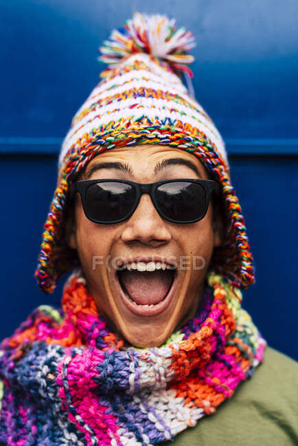 Счастливый подросток с разноцветной вязаной шляпой и шарфом перед синей стеной — стоковое фото