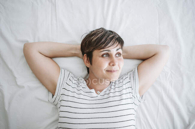 Задумчивая женщина с руками за головой лежит на кровати дома — стоковое фото