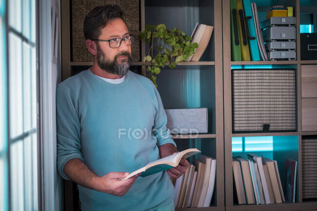 Homme d'affaires réfléchi tenant livre tout en s'appuyant sur la bibliothèque à la maison — Photo de stock