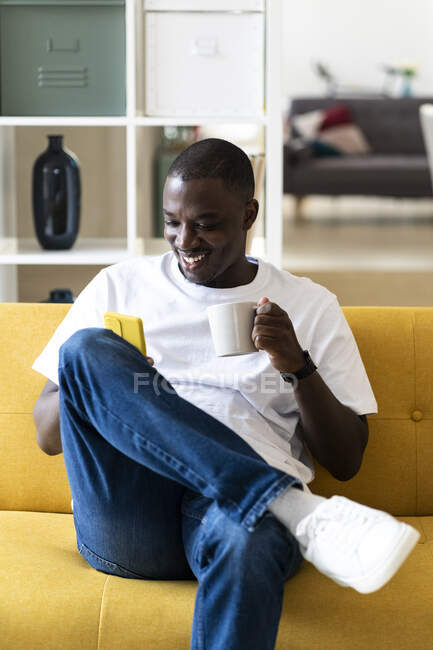 Homme tenant tasse à l'aide d'un téléphone intelligent tout en étant assis sur le canapé à la maison — Photo de stock