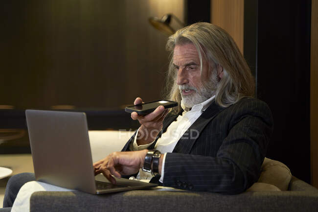 Mature businessman sending voicemail through smart phone while using laptop at hotel — Stock Photo