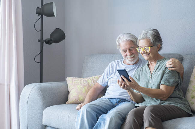 Улыбающаяся пожилая женщина делит мобильный телефон с мужчиной на диване — стоковое фото