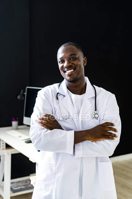 Médico varón sonriente con bata de laboratorio de pie con los brazos cruzados en la clínica médica - foto de stock