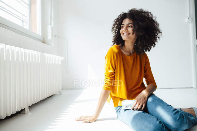 Mujer afro sonriente con teléfono móvil sentado en el suelo en casa - foto de stock