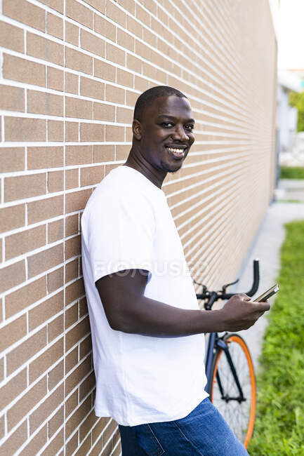 Sonriente hombre adulto medio con teléfono inteligente de pie por la pared de ladrillo - foto de stock