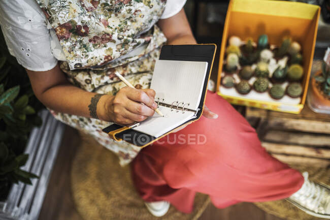 Floristería femenina madura escribiendo en libro mientras está sentada en una floristería - foto de stock