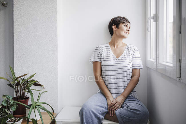 Woman looking through window while sitting by plants at home — Stock Photo