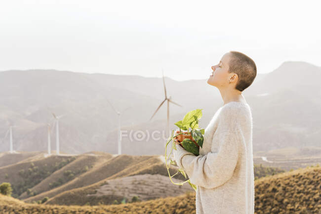 Жінка з закритими очима тримає горщик рослини на вітроелектростанції в сонячний день — стокове фото