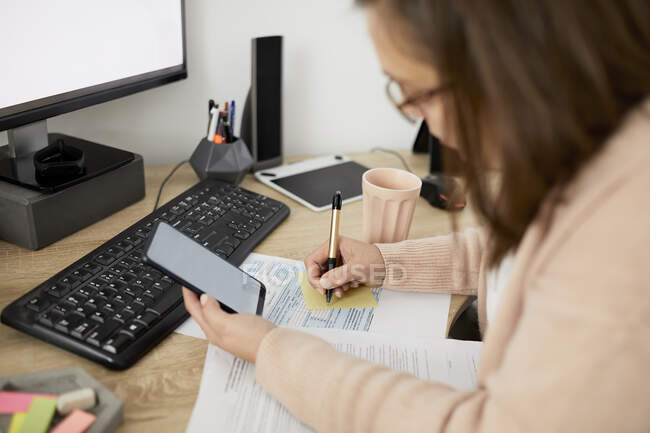 Femme d'affaires tenant un téléphone intelligent et écrivant assis au bureau — Photo de stock