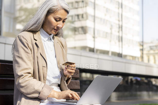 Woman doing online payment with credit card while using laptop on bench — Stock Photo
