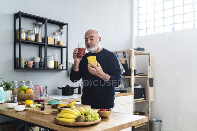 Старший чоловік з розумним телефоном п'є коктейль вдома. — стокове фото