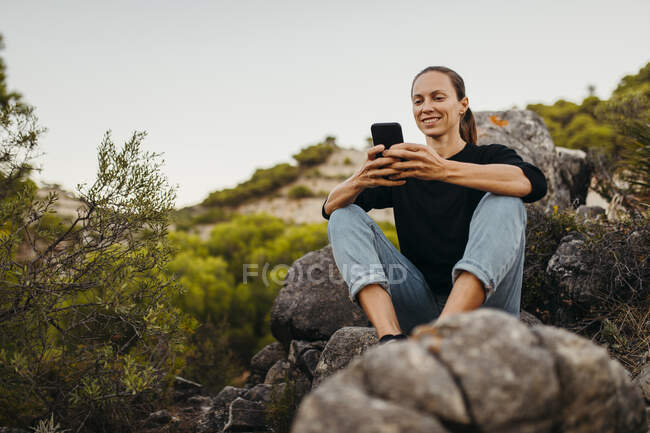 Mujer sonriente usando el teléfono móvil en la roca - foto de stock