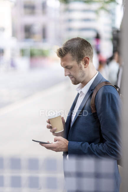 Male professional holding disposable cup using mobile phone at bus stop — Stock Photo
