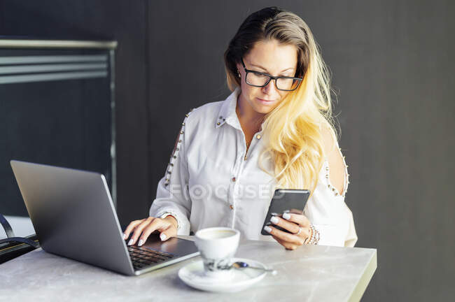 Mujer de negocios rubia usando el teléfono móvil mientras trabaja en la cafetería - foto de stock