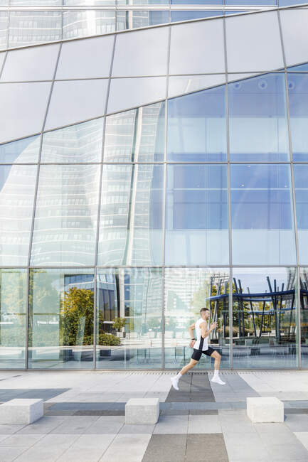 Young male athlete running on footpath by glass building — Stock Photo