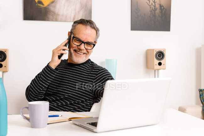 Heureux homme mature parlant sur téléphone intelligent tout en utilisant un ordinateur portable dans le bureau à la maison — Photo de stock