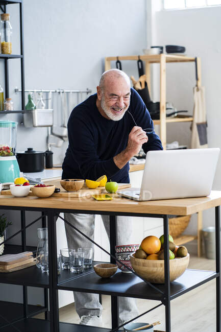 Allegro uomo anziano utilizzando il computer portatile al bancone della cucina — Foto stock
