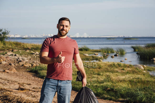 Sorridente attivista maschio gesticolando pollici in su mentre in piedi sulla riva del fiume durante la giornata di sole — Foto stock