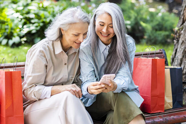 Maduras amigas con bolsas de compras compartiendo el teléfono inteligente mientras están sentadas en el banco - foto de stock