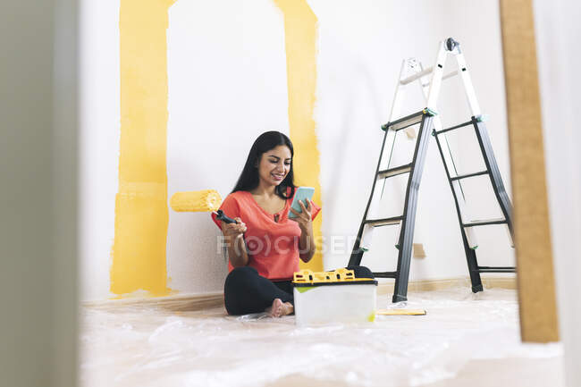 Sorrindo jovem com rolo de pintura usando telefone inteligente na sala de estar — Fotografia de Stock