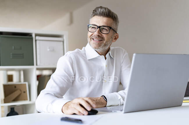Hombre de negocios sonriente con portátil contemplando en la oficina - foto de stock