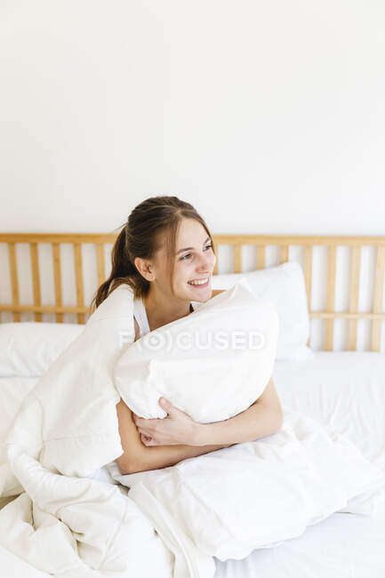 Smiling young woman embracing pillow while sitting on bed in bedroom — Stock Photo