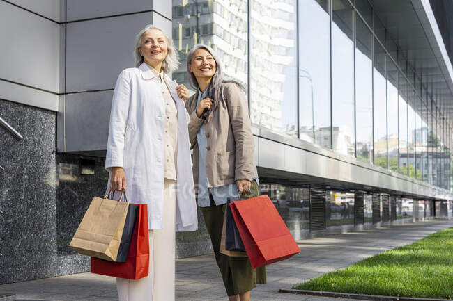 Mulheres sorrindo olhando para longe enquanto segurando sacos de compras fora do shopping — Fotografia de Stock