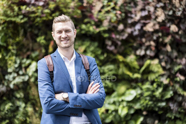 Empresário sorridente de pé com os braços cruzados na frente da planta verde — Fotografia de Stock