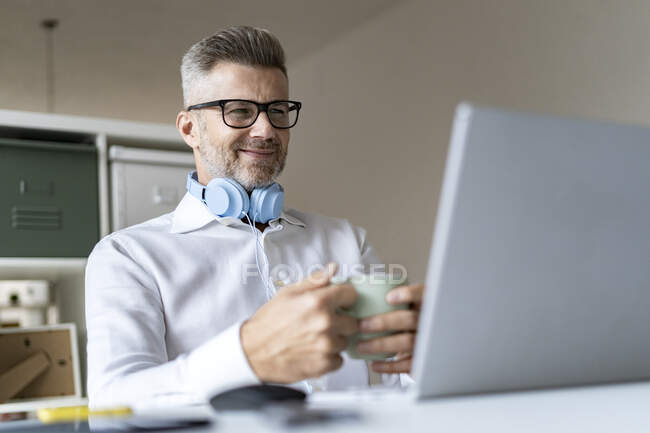 Усміхнений бізнесмен з чашкою кави на відеоконференції через ноутбук у офісі — стокове фото