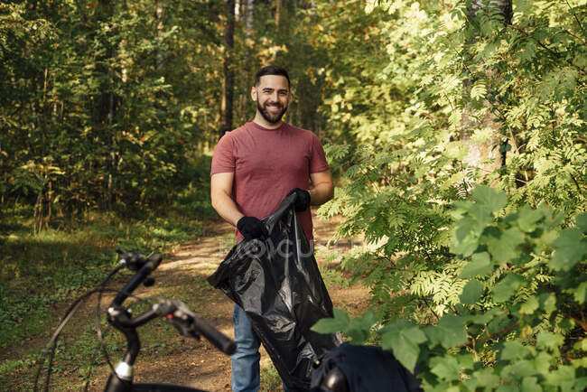 Sorridente volontario maschio che tiene il sacco della spazzatura nella foresta — Foto stock