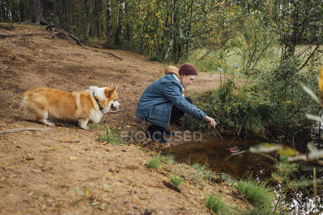 Menino mergulhando vara na água enquanto se agacha perto do lago na floresta — Fotografia de Stock