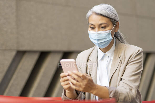 Mature woman wearing protective face mask using smart phone at sidewalk cafe — Stock Photo