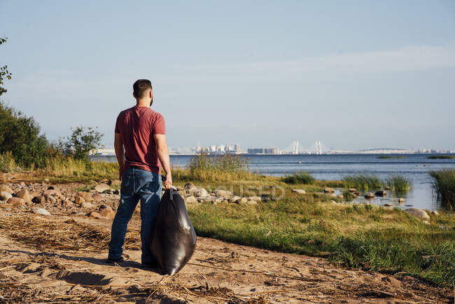 Hombre medio adulto voluntario con bolsa de basura mirando el río - foto de stock
