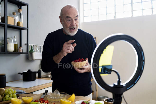 Senior man with fruit bowl vlogging through smart home in kitchen — Stock Photo