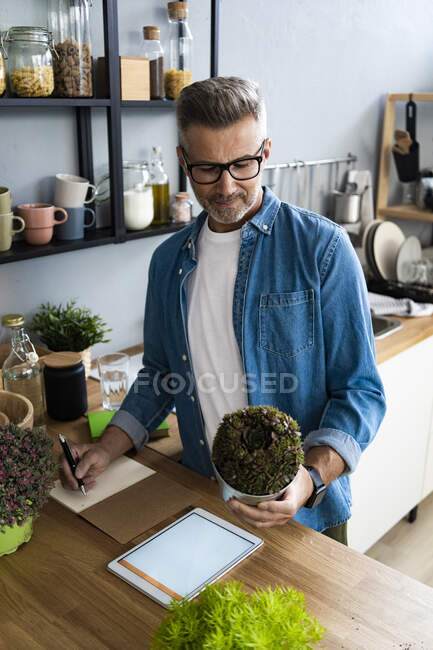 Man checking plant while writing on paper at kitchen counter — Stock Photo