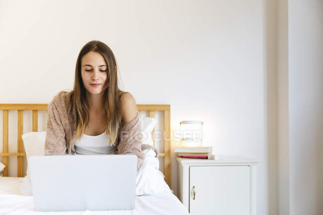 Jovem mulher usando laptop enquanto se senta na cama em casa — Fotografia de Stock