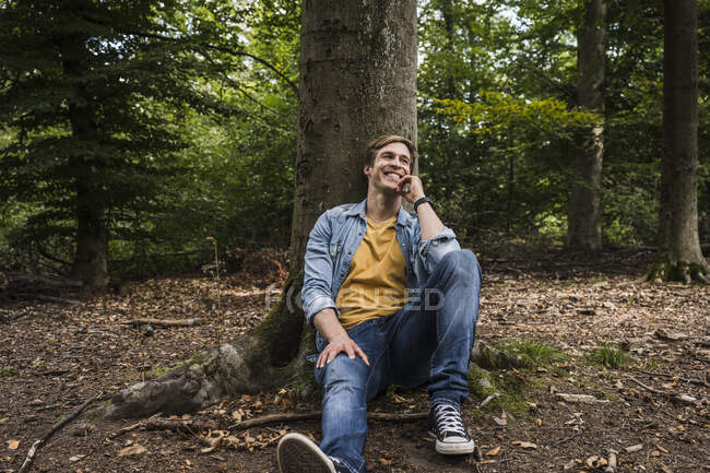 Щасливий чоловік з рукою на підборідді сидить біля стовбура дерева в лісі. — стокове фото