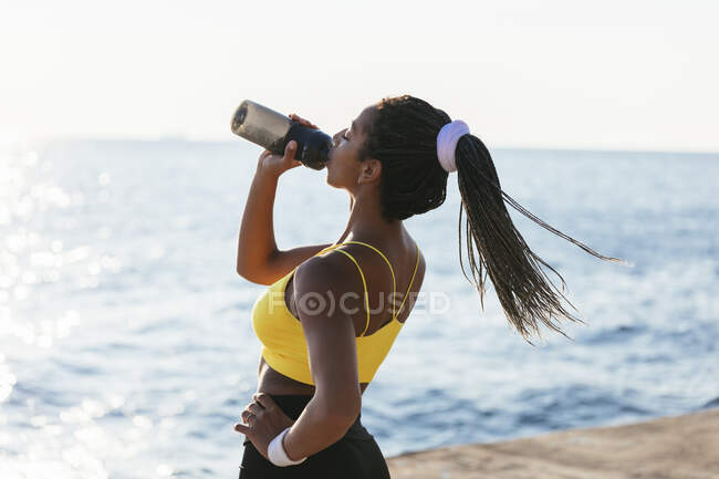Молодая спортсменка пьет воду, стоя у моря — стоковое фото