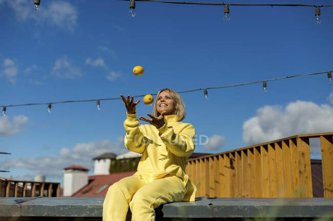 Sorridente donna giocoleria limoni mentre seduto sul muro di contenimento — Foto stock