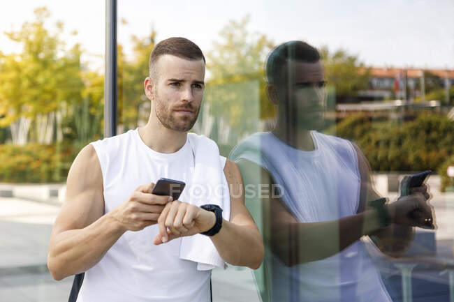 Deportista con teléfono móvil y reloj de pulsera apoyado en la pared de cristal - foto de stock