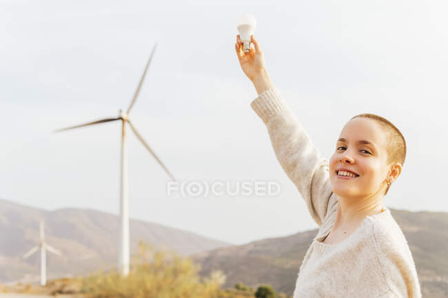 Mulher feliz com mão levantada segurando lâmpada no parque eólico — Fotografia de Stock