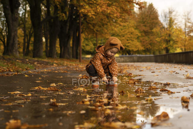 Girl wearing brown cap and raincoat crouching on road in park — Stock Photo