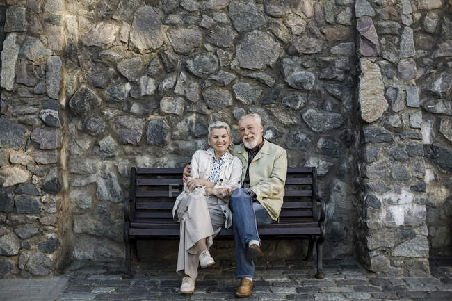 Couple souriant assis avec les jambes croisées au genou devant un mur de pierre — Photo de stock