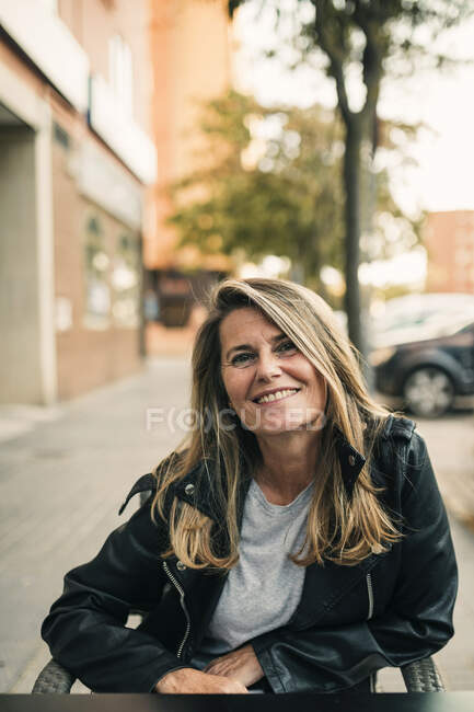 Smiling blond woman wearing leather jacket while sitting at sidewalk cafe — Stock Photo