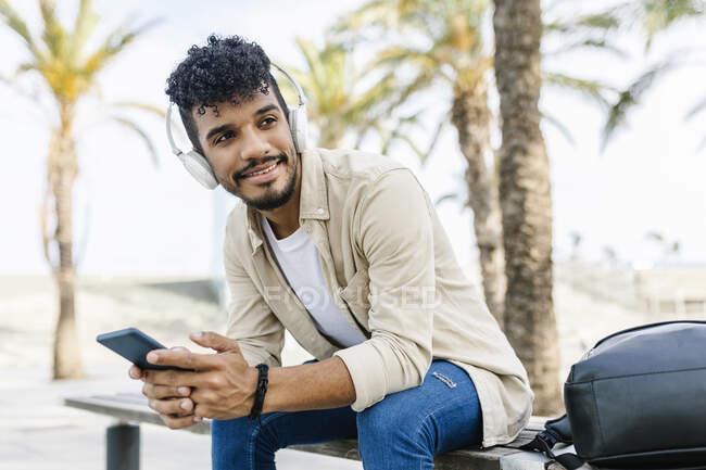 Young man wearing wireless headphones holding mobile phone while sitting on bench — Stock Photo
