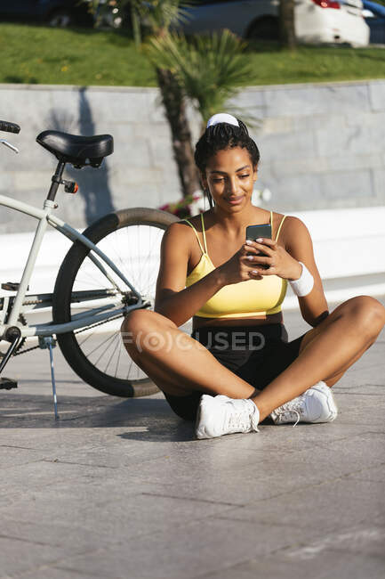 Jeune sportive utilisant un téléphone intelligent assis à vélo sur un sentier piétonnier — Photo de stock