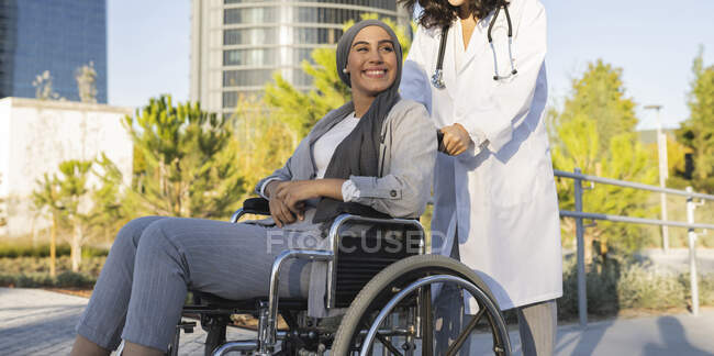 Жінка-лікар штовхає інвалідне крісло усміхненої інвалідної жінки на пішохідному шляху — стокове фото